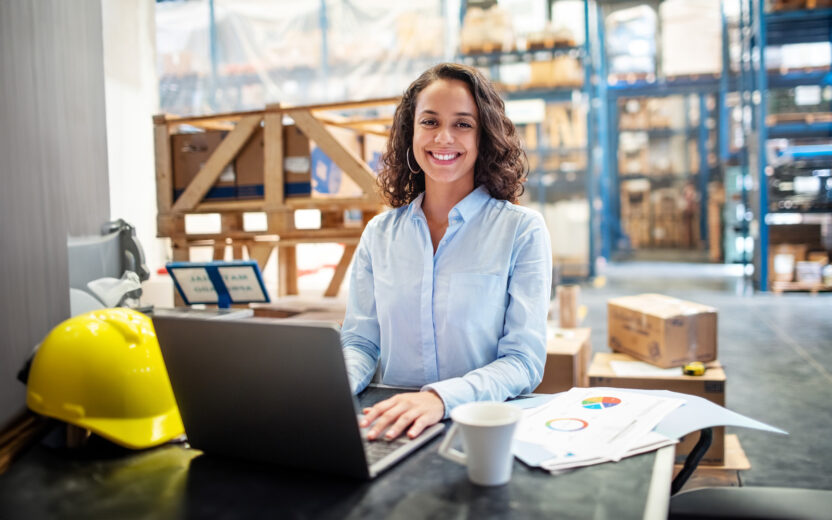 Smiling business woman working on laptop at a warehouse. Businesswoman with a laptop working at a distribution warehouse.