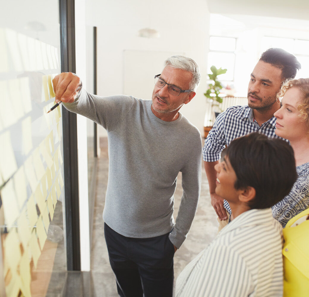 Mature businessman and his diverse team brainstorming with yellow adhesive notes on a glass wall in a bright modern office