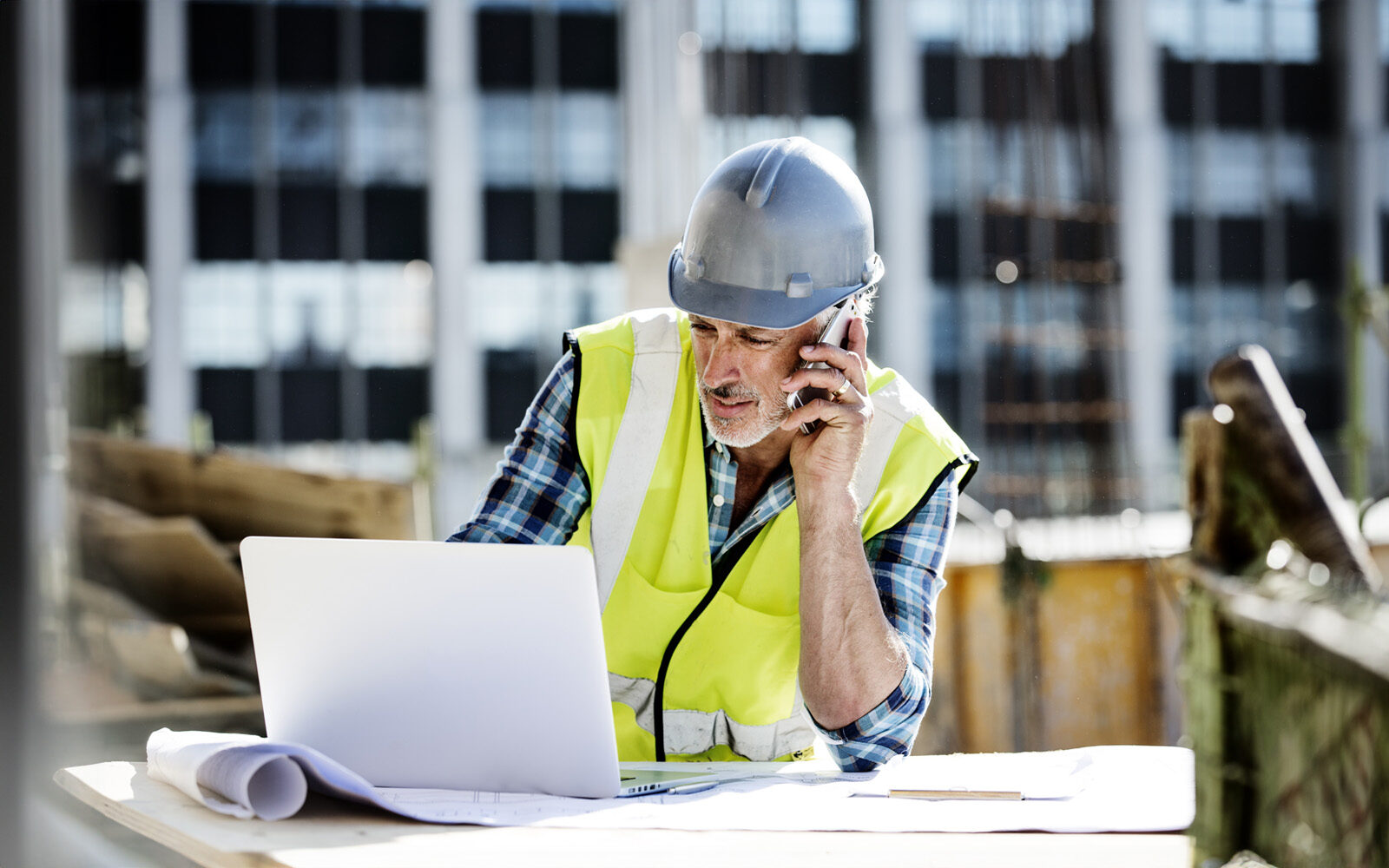 Male architect using mobile phone and laptop at construction site for digital resource planning.