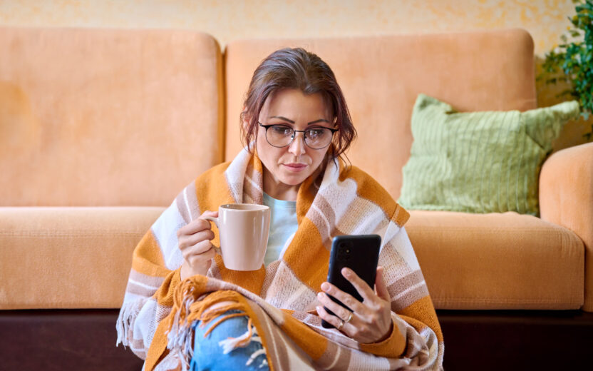 The season of cold weather is autumn winter, a woman under a woolen warm blanket with a cup of hot tea using a smartphone. Middle-aged female resting relaxing at home