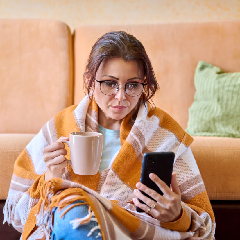 The season of cold weather is autumn winter, a woman under a woolen warm blanket with a cup of hot tea using a smartphone. Middle-aged female resting relaxing at home