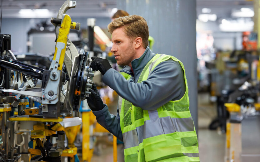 Confident mid adult engineer examining car chassis at automobile industry. Handsome male supervisor is working on car part in factory. He is wearing reflective clothing.