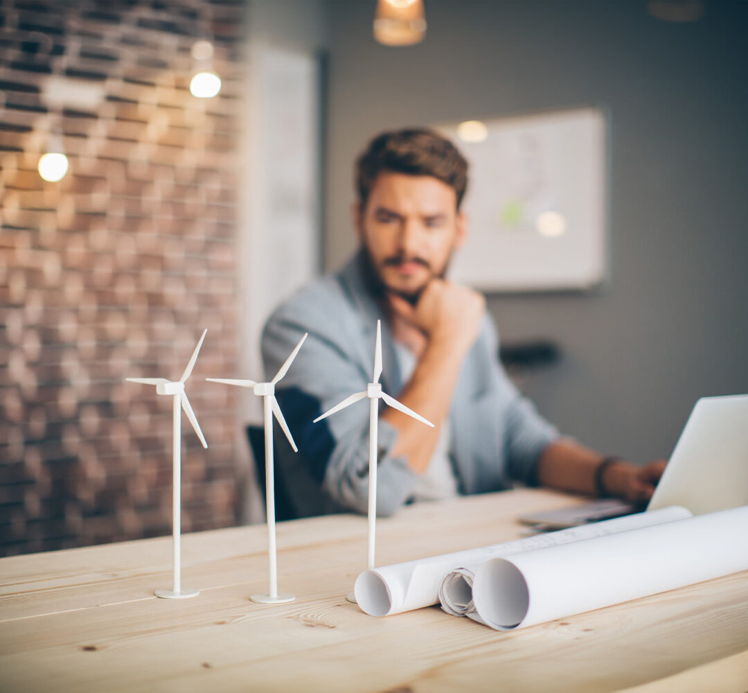 Young engineer at modern office space, sitting at the table and working on energy project. Focus on wind turbine models on the table.