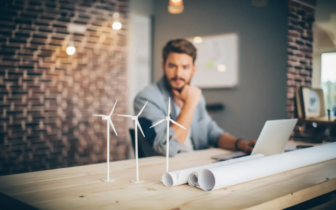 Young engineer at modern office space, sitting at the table and working on energy project. Focus on wind turbine models on the table.