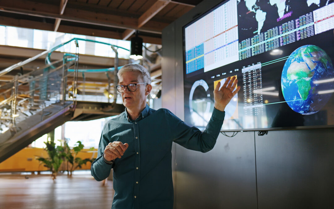 Image of a mature man conducting a seminar with the aid of a large screen displaying graphs and data associated with images of the earth