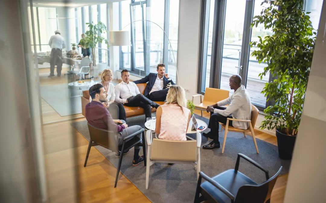 Fotografia de perspectiva elevada sob um grupo de pessoas, tirada através da janela, que mostra colegas de negócios num momento descontraído, a conversar no lobby de um escritório moderno.