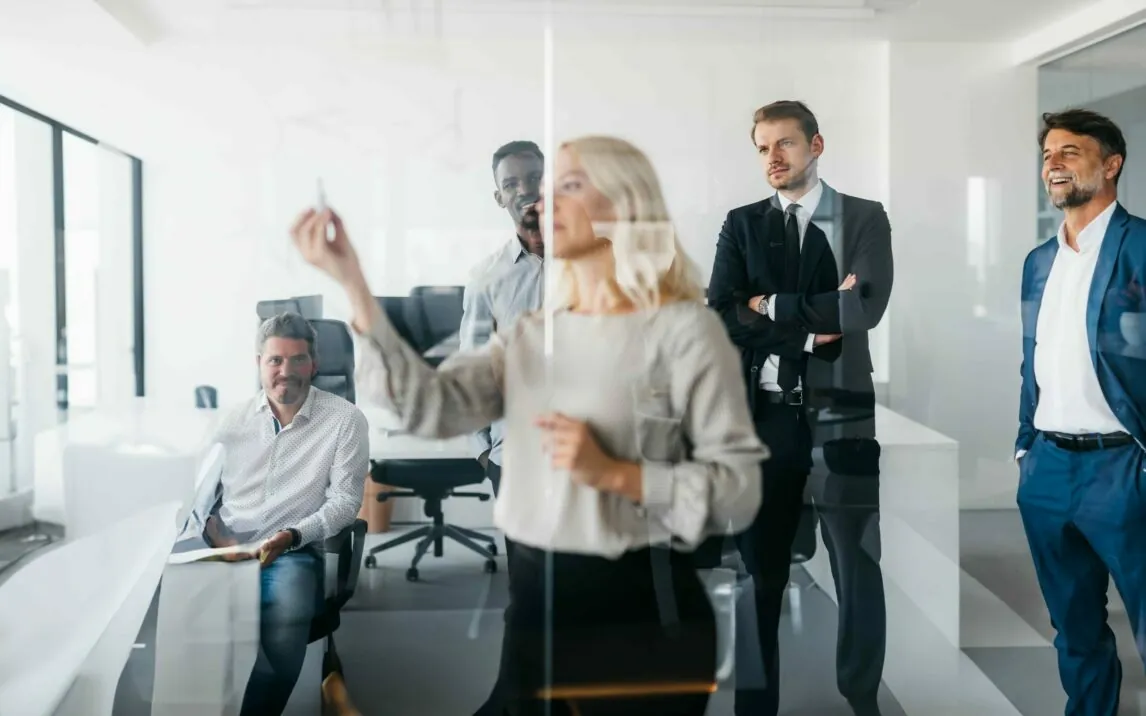 A business woman is writing on a glass wall, while her colleagues are listening to her. They are smartly dressed and professional. The office is bright and spacious. Horizontal daylight indoor photo.