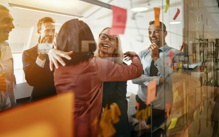 Two smiling young businesswomen hugging while brainstorming with sticky notes on a glass wall with colleagues in a modern office