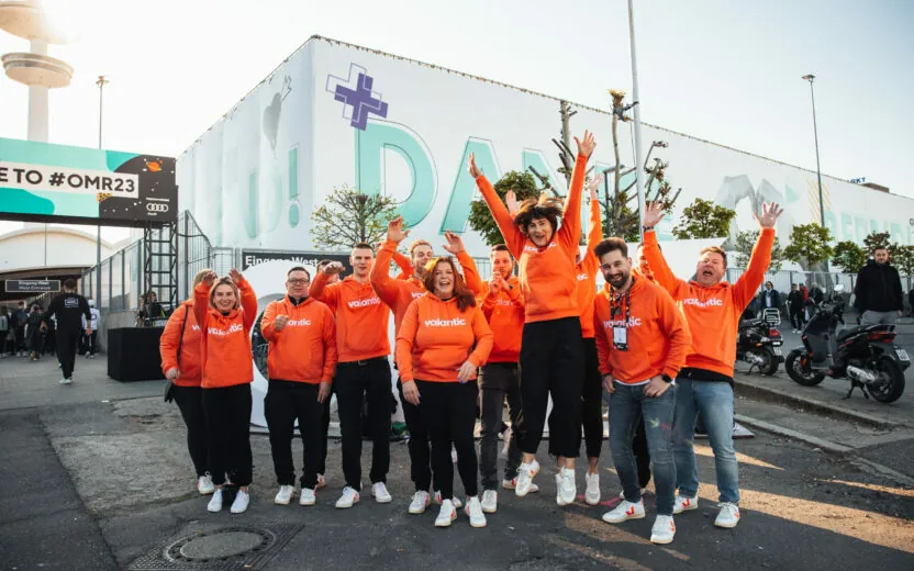 Eine Gruppe von Menschen in passenden orangefarbenen Kapuzenpullovern posiert voller Energie vor einem großen Veranstaltungsgebäude. Einige springen vor Aufregung in die Luft.