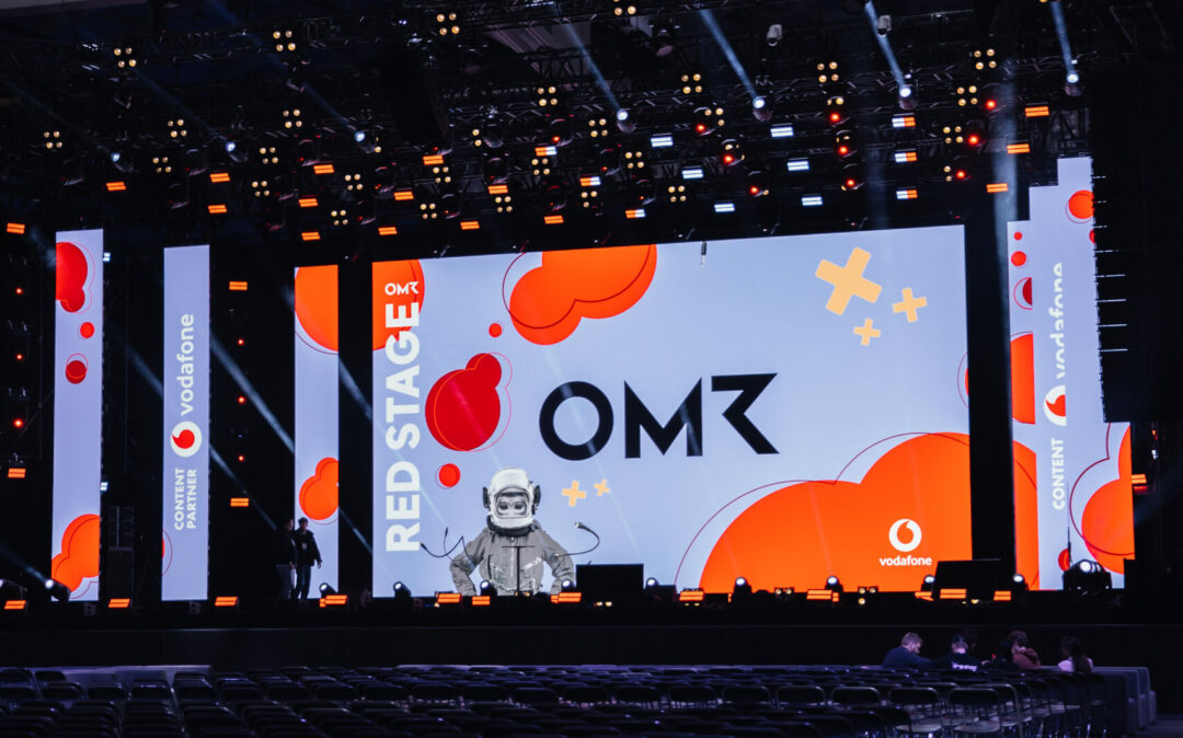 Eine große Veranstaltungsbühne mit einem Bildschirm, auf dem die Logos „RED STAGE“ und „OMR“ unter bunten Grafiken zu sehen sind. Reihen leerer Stühle füllen den Vordergrund. Darüber sind Deckenleuchten und -leuchten zu sehen.