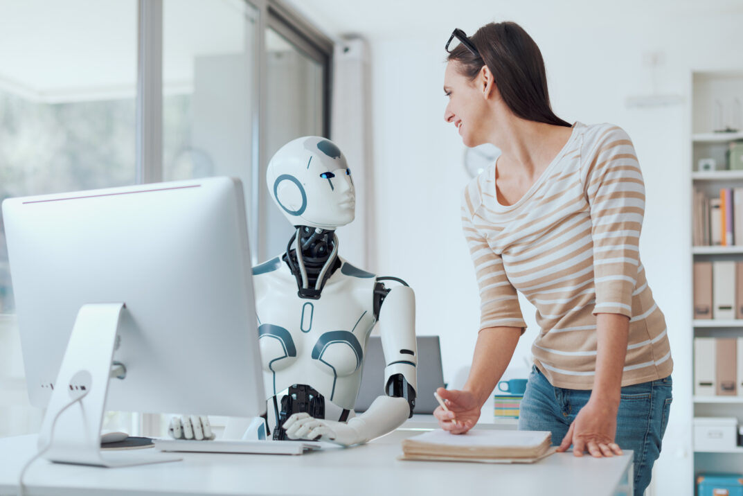 Eine Frau lächelt, während sie KI-Beratung mit einem humanoiden Roboter durchführt, der an einem Bürotisch neben einem Computermonitor sitzt.