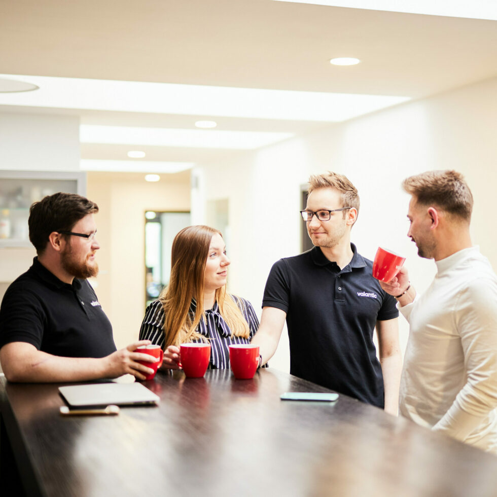 Four people standing at a counter, holding red mugs and engaging in conversation in a brightly lit room. One person is wearing a white shirt, and the others are in dark attire. A laptop is on the counter.