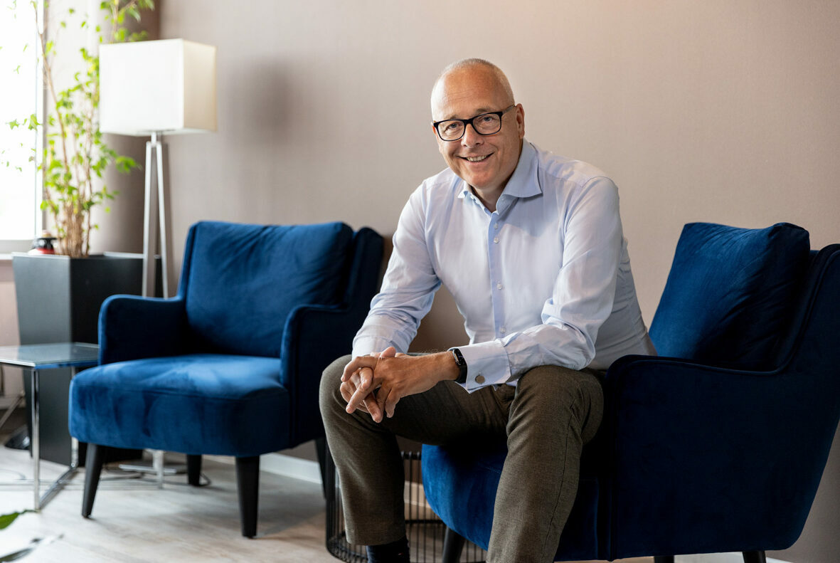 A man in glasses and a light blue shirt is sitting on a blue chair in a modern room with another blue chair, a lamp, and a potted plant.