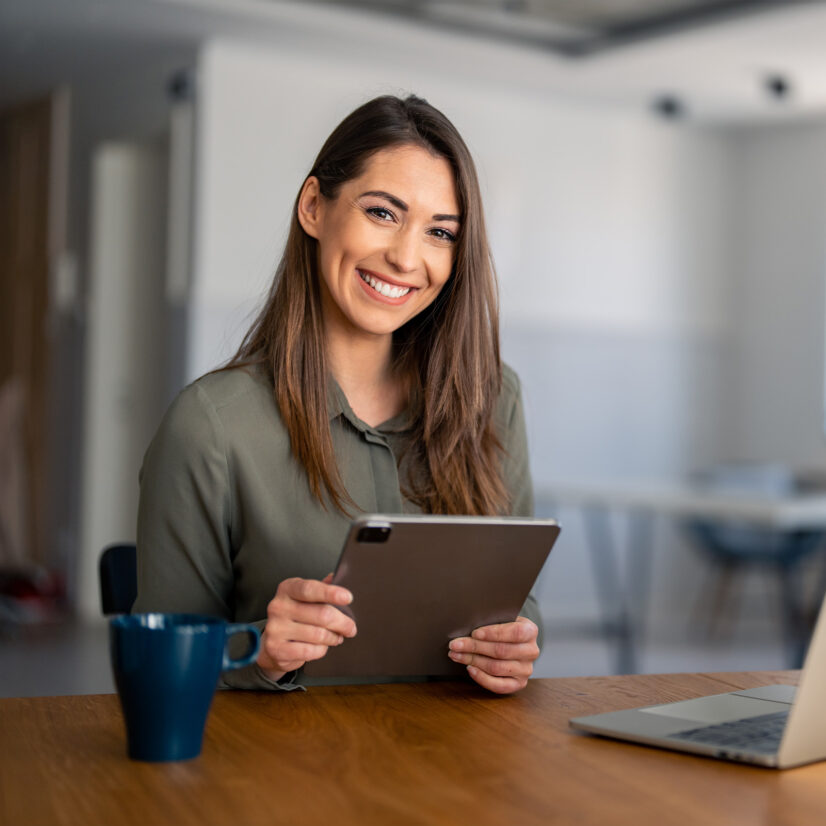 Porträt einer lächelnden, erfüllten jungen Frau, die ein digitales Tablet-Gerät in der Hand hält, in die Kamera blickt und zu Hause an einem Schreibtisch sitzt, vor sich einen Laptop und eine Tasse Kaffee.