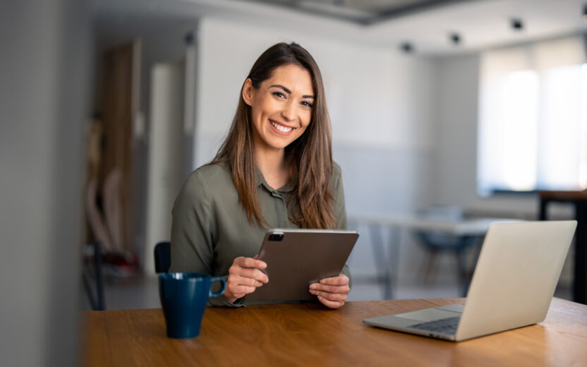 Portrait of smiling fulfilled young woman holding digital tablet device, looking at camera, sitting at desk at home with laptop computer in front of her and cup of coffee.