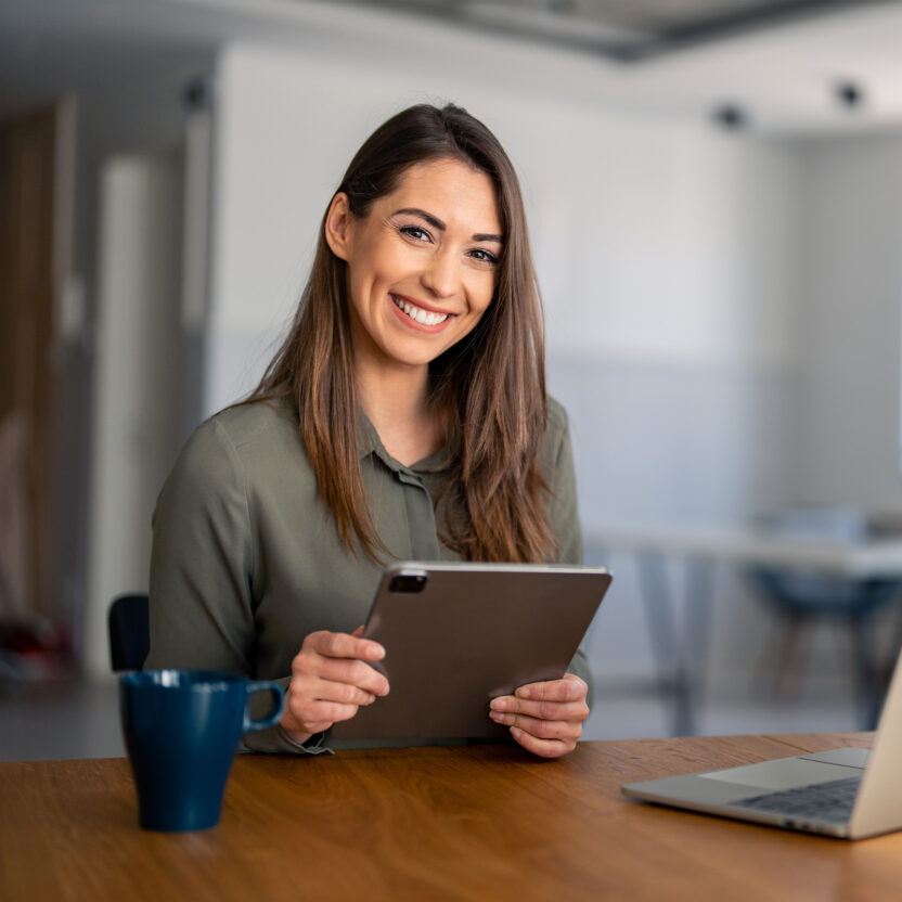 Portrait of smiling fulfilled young woman holding digital tablet device, looking at camera, sitting at desk at home with laptop computer in front of her and cup of coffee.