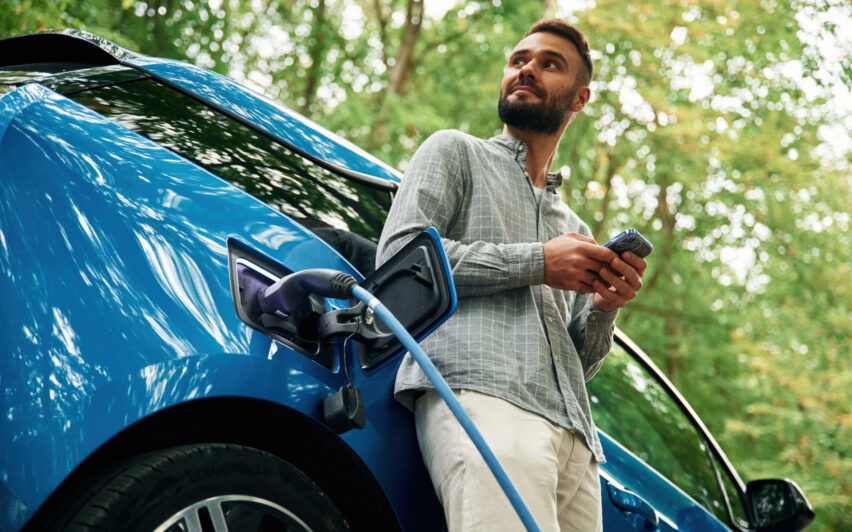 Trees behind. Handsome man is standing outdoors near electric automobile and waiting for charging.