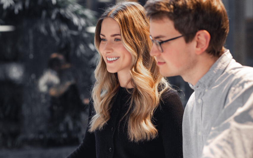 Eine lächelnde junge Frau mit langen blonden Haaren und ein junger Mann mit Brille und weißem Hemd im Profil.