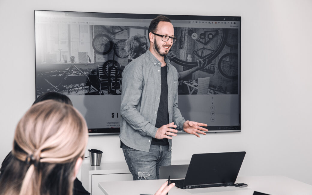 Ein Mann steht vor einem Bildschirm und hält in einem modernen Konferenzraum eine Präsentation vor zwei sitzenden Personen.