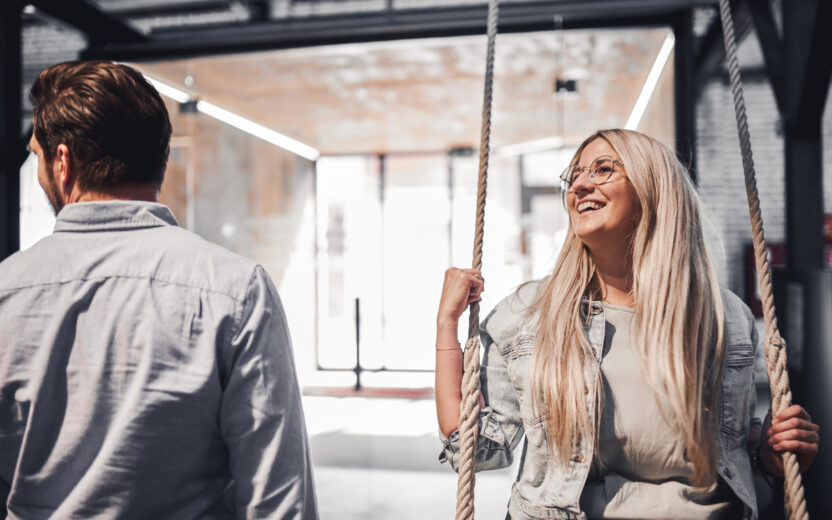 Foto von einer jungen Dame mit blonden Haaren, die auf einer Schaukel in einem Büro sitzt. Neben ihr sieht man einen jungen Mann von hinten.
