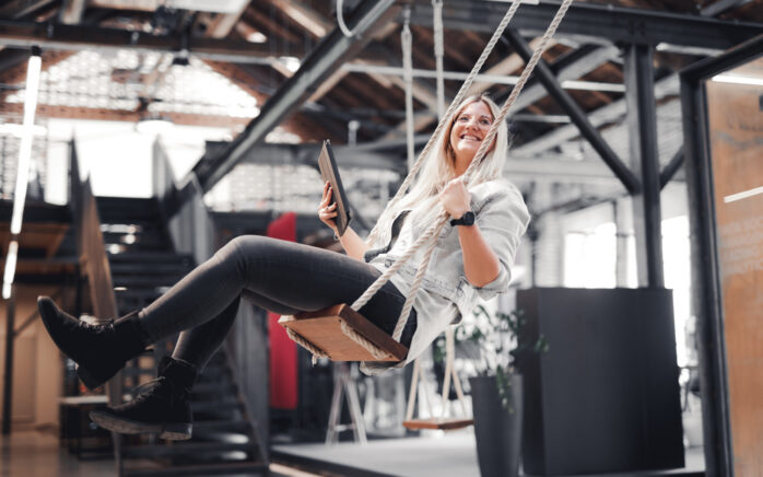 Eine Person sitzt in einem modernen Büroraum im Industriestil auf einer Schaukel, lächelt und hält ein Tablet.