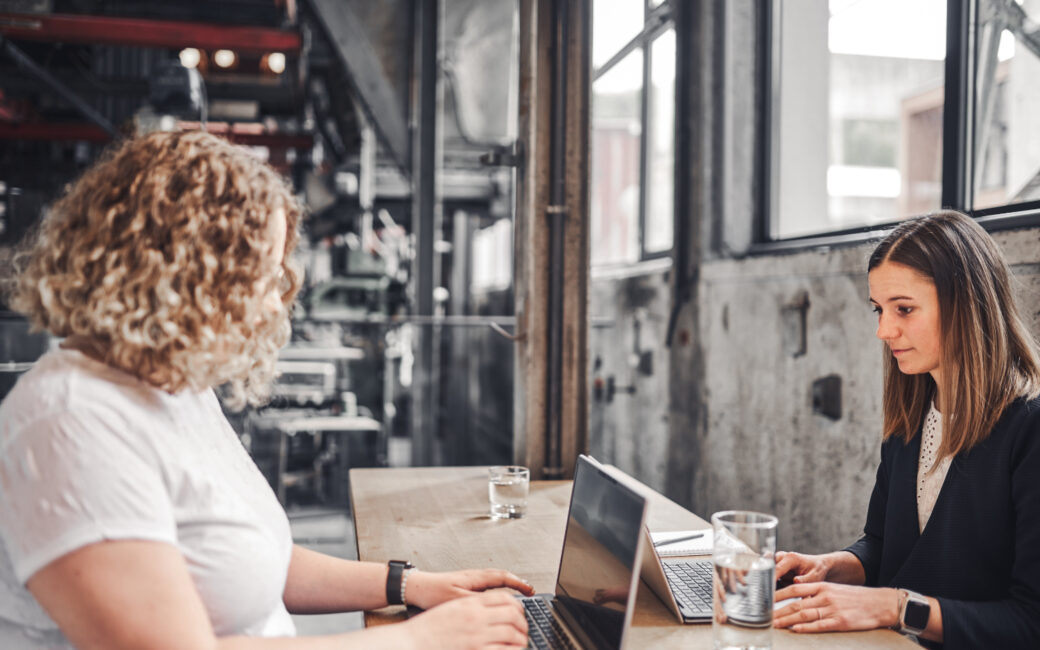 In einem im Industriestil eingerichteten Raum sitzen sich zwei Frauen an einem Holztisch gegenüber. Jede von ihnen nutzt einen Laptop, vor ihnen stehen Gläser mit Wasser.