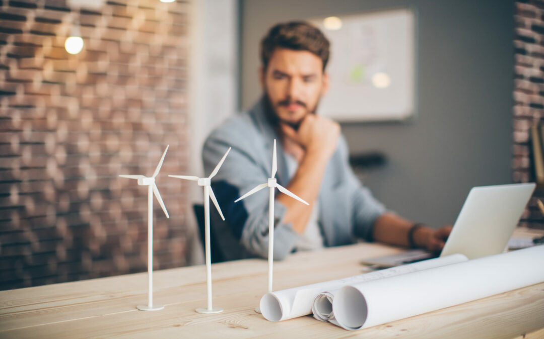 Junger Ingenieur in einem modernen Büro, der am Tisch sitzt und an einem Energieprojekt arbeitet. Fokus auf Windkraftanlage Modelle auf dem Tisch.