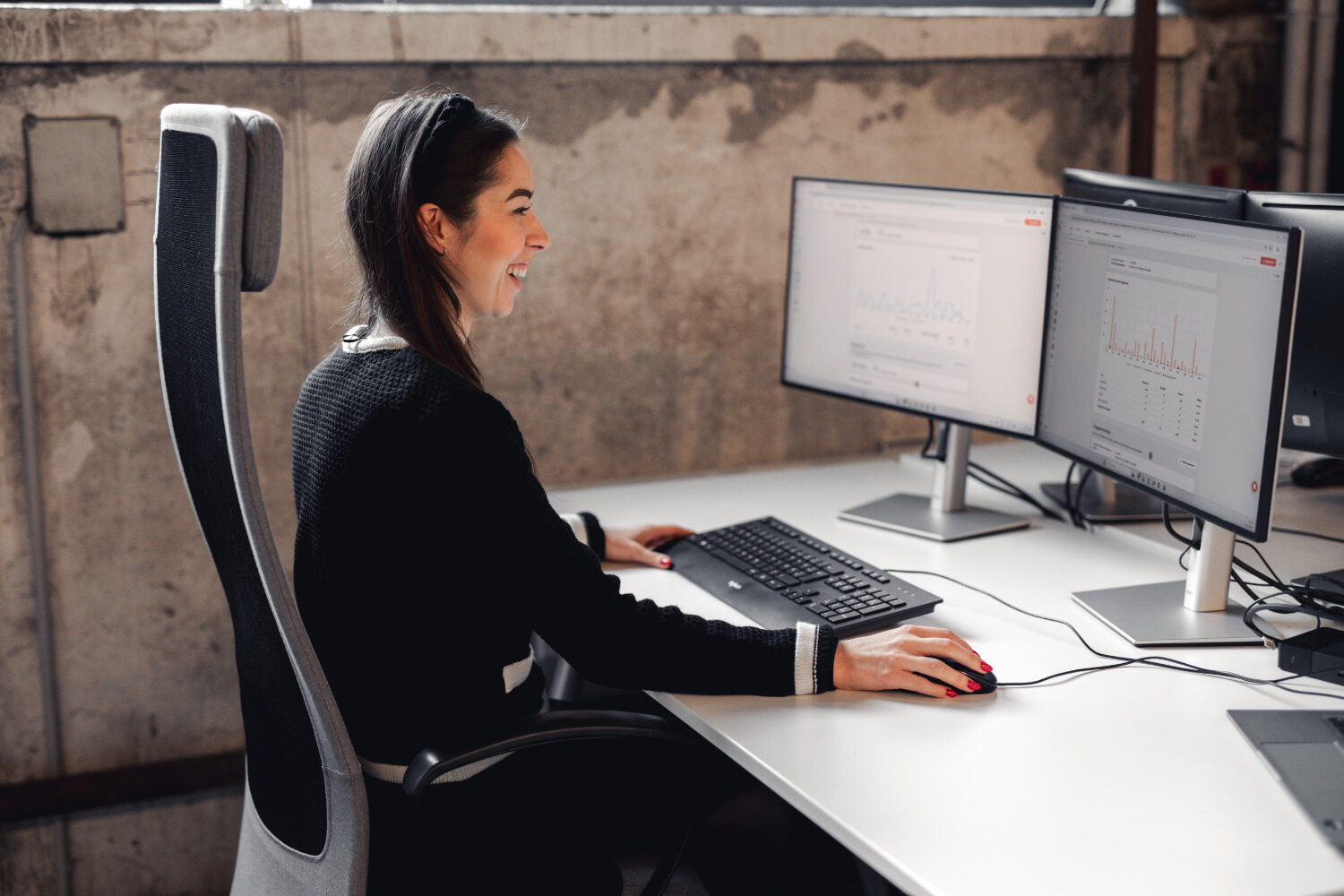 Eine junge lächelnde Frau mit dunkler Kleidung sitzt an ihre Arbeitsplatz.