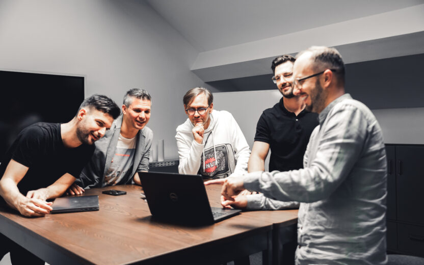 Fünf Männer stehen in einem Meetingraum zusammen und besprechen etwas am Laptop.