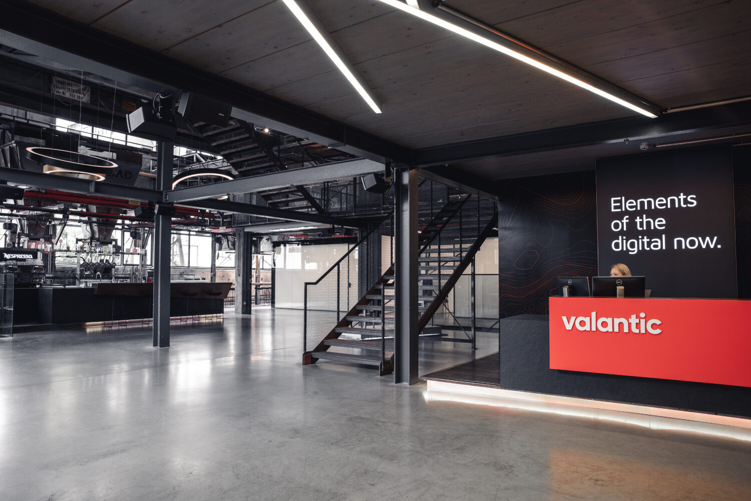 Das Bild zeigt das Foyer des valantic-Büros in Salzburg. Das Office ist im Industrial Style, hat einen orangenen Empfangstresen mit dem valantic-Logo, eine Treppe aus Eisen, eine moderne Bar und Meeting-Räume mit Milchglas.