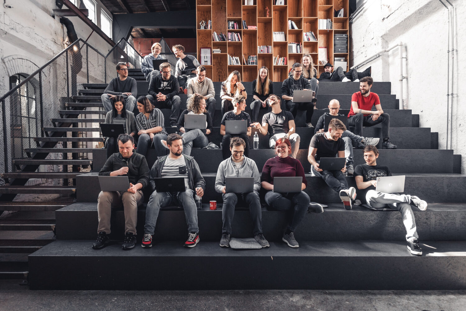Eine Gruppe aus Frauen und Männern sitzt mit ihren Laptops auf einer dunklen Tribüne in einem Vortrags-Saal. Sie arbeiten, unterhalten sich oder lachen.