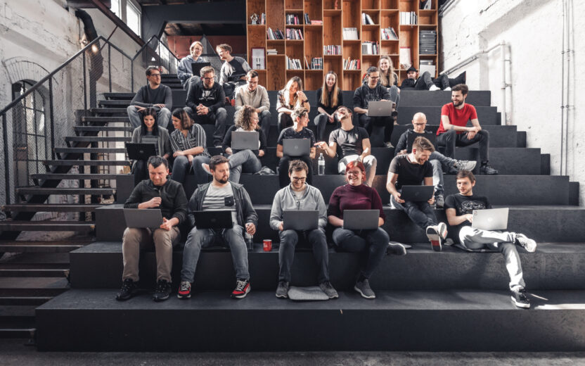 Eine Gruppe aus Frauen und Männern sitzt mit ihren Laptops auf einer dunklen Tribüne in einem Vortrags-Saal. Sie arbeiten, unterhalten sich oder lachen.