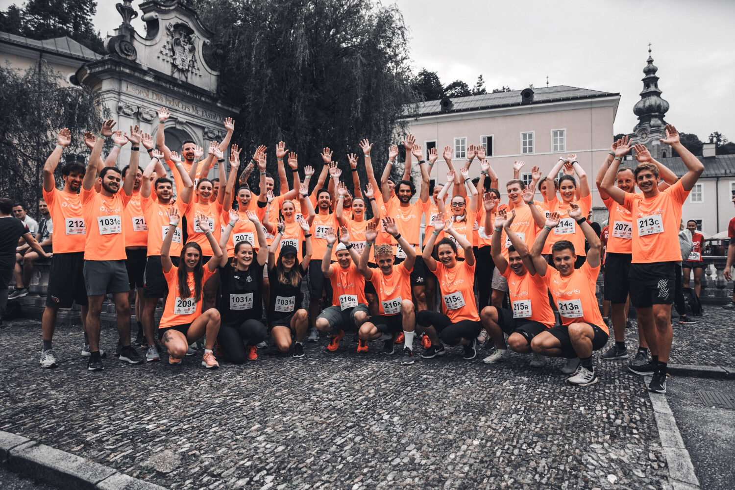 Eine große Gruppe von elements-Mitarbeiter*innen jubeln vor dem Domplatz Salzburg. Sie tragen alle orange Sportshirts mit dem elements-Logo.