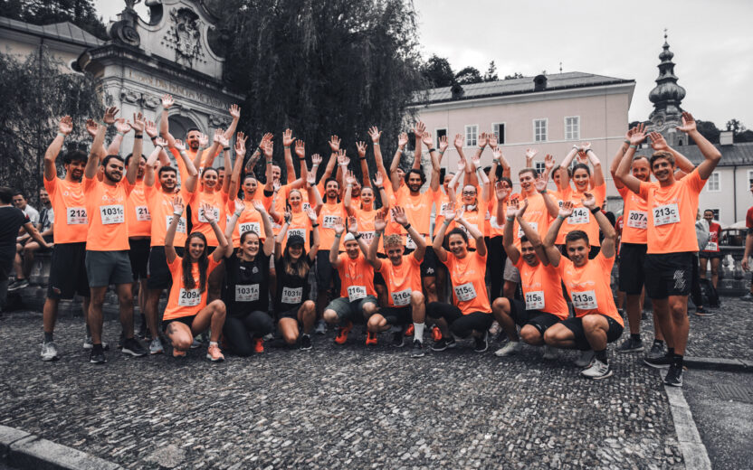Eine große Gruppe von elements-Mitarbeiter*innen jubeln vor dem Domplatz Salzburg. Sie tragen alle orange Sportshirts mit dem elements-Logo.