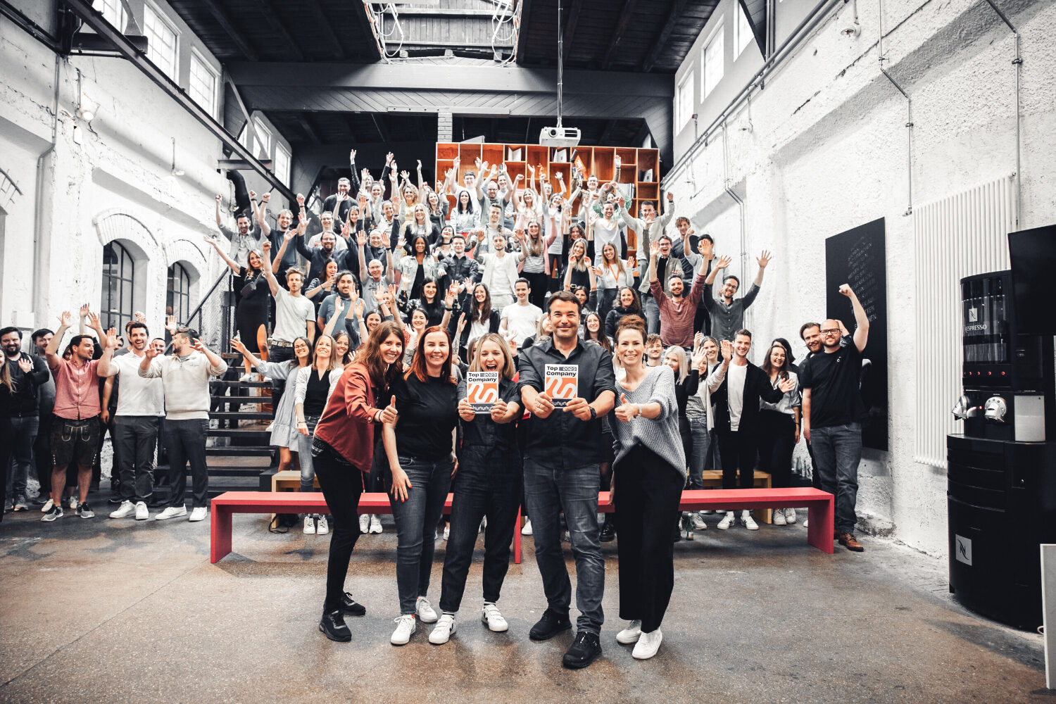 Aufnahme von einer großen Gruppe elements-Mitarbeitende, die auf einer Treppe stehen und jublen. Ganz vorne im Bild stehen 5 Mitarbeiter*innen von elements und halten die kununu-Top-Company-Auszeichnung ins Bild.
