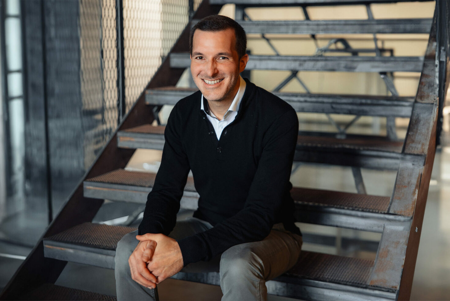 A man in a black sweater and khaki pants sits on a metal staircase indoors, smiling at the camera.