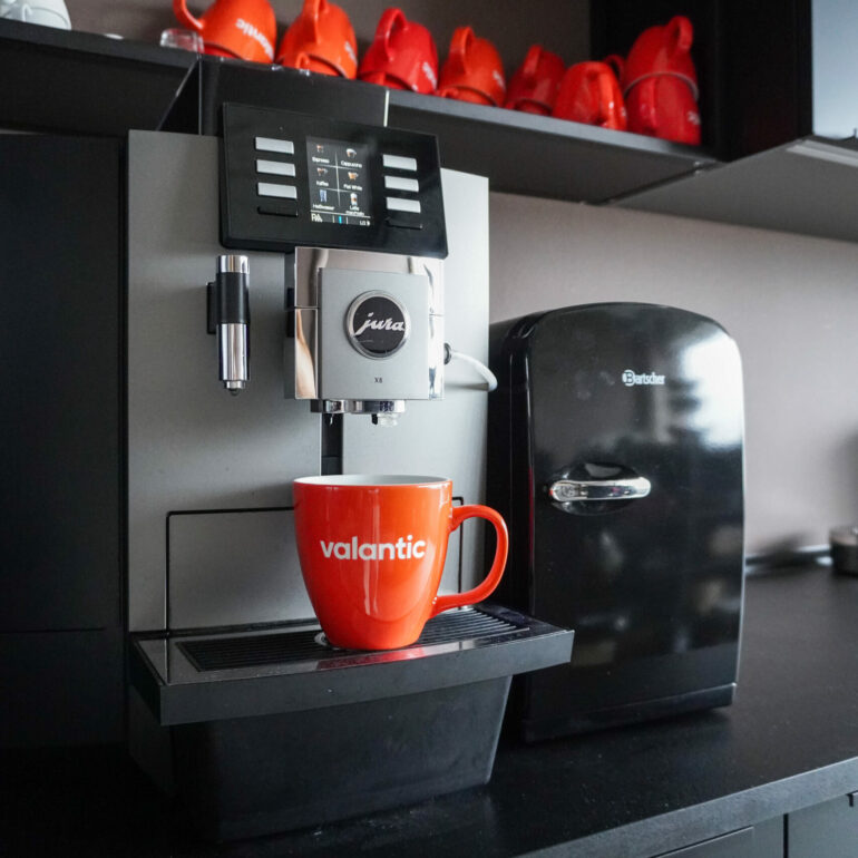 Eine Kaffeemaschine gibt Kaffee in einen roten Becher mit der Aufschrift „Valantic“ auf einer Arbeitsplatte aus. Darüber stehen rote und weiße Tassen auf Regalen und rechts davon steht ein kleiner schwarzer Kühlschrank.