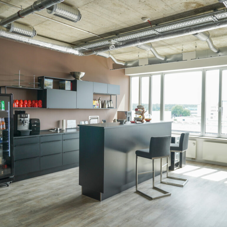 Dunkle Küche mit Theke, zwei Barhockern, einer Kaffeemaschine und langer Fensterfront im Hintergrund