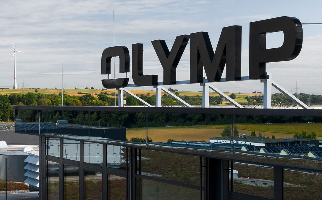 Außenansicht eines Gebäudes mit einem großen „OLYMP“-Schild auf dem Dach, vor einem klaren blauen Himmel mit flauschigen Wolken.