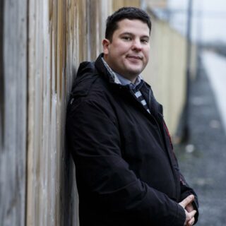 Was wäre, wenn ein Mann in einer dunklen Jacke auf einem gepflasterten Weg an einem Holzzaun lehnt und nachdenklich in die Kamera blickt?