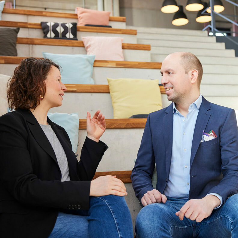 Two people sit and talk on tiered seating steps with pillows behind them. Both are dressed in business casual attire.