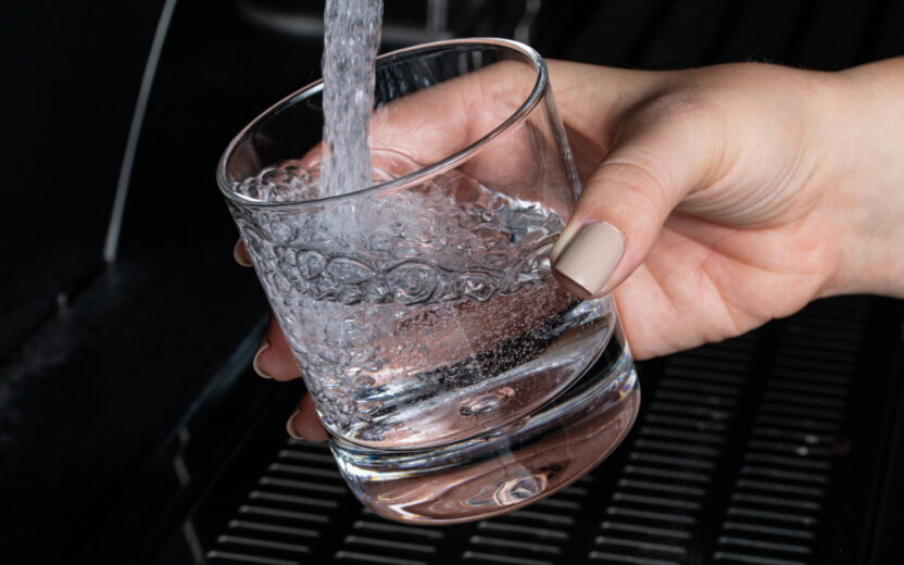 Nahaufnahme von einem Glas, in das frisches Wasser aus einem Wasserhahn läuft. Das Glas wird von einer Frauenhand festgehalten. Der Hintergrund ist schwarz.