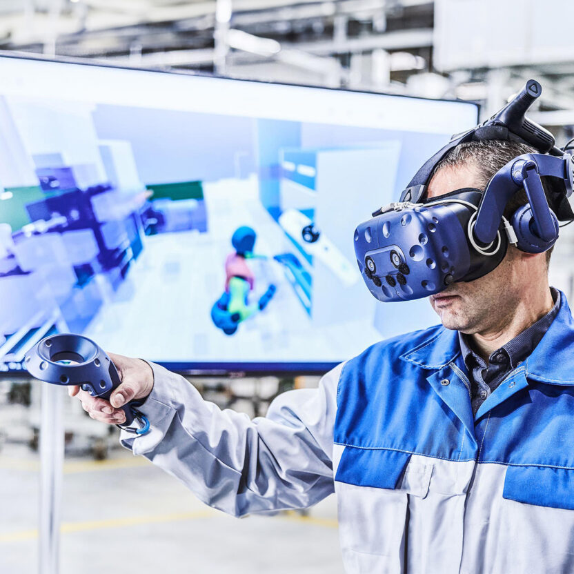A man in a blue-grey work jacket is wearing VR goggles and holding a remote control. In the background, you can see a blurred screen showing the VR program.
