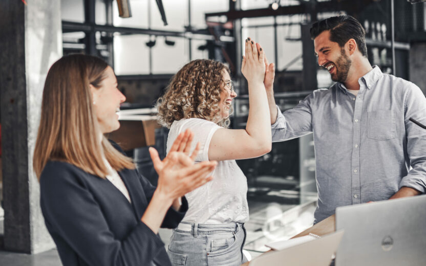 Three colleagues celebrate success in a modern office environment. Two people give each other a high-five while another person claps nearby.