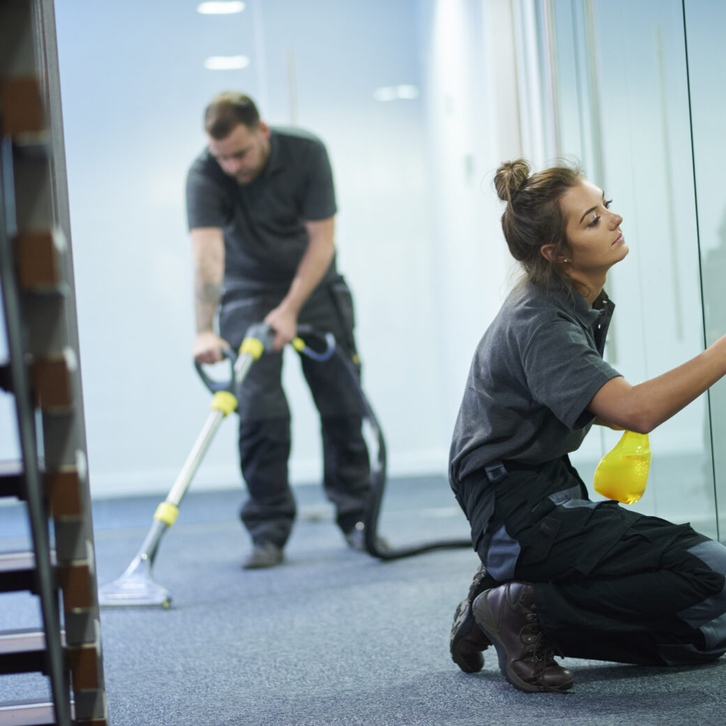 a female cleaning contractor is polishing the glass partition offices whilst In the background a male colleague steam cleans an office carpet in a empty office in between tenants. The female is smiling to camera.