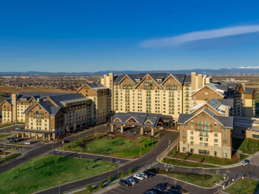 Aerial view of a large resort complex hosting a Visionary Global Leadership seminar, with multiple buildings surrounded by parking lots and greenery, set against a backdrop of mountains and a blue sky.