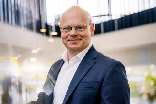 A person in a suit and glasses stands indoors, smiling. Background features modern architecture with natural light.