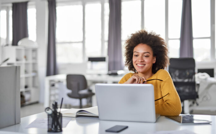 A person in a yellow sweater smiles while looking at a laptop in a bright office. Office supplies and a phone are on the desk.
