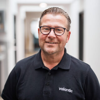 Ein Mann mit Brille und schwarzem Poloshirt mit dem „valantic“-Logo steht in einem hellen Flur und scheint mit Kollegen von ORCA Software Produktdatenstrategien zu besprechen.