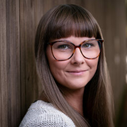 A woman with long brown hair and glasses leans against a wooden wall, her smile as warm as her white sweater. She seems like someone you could chat with for hours, almost like a friendly chatbot brought to life.
